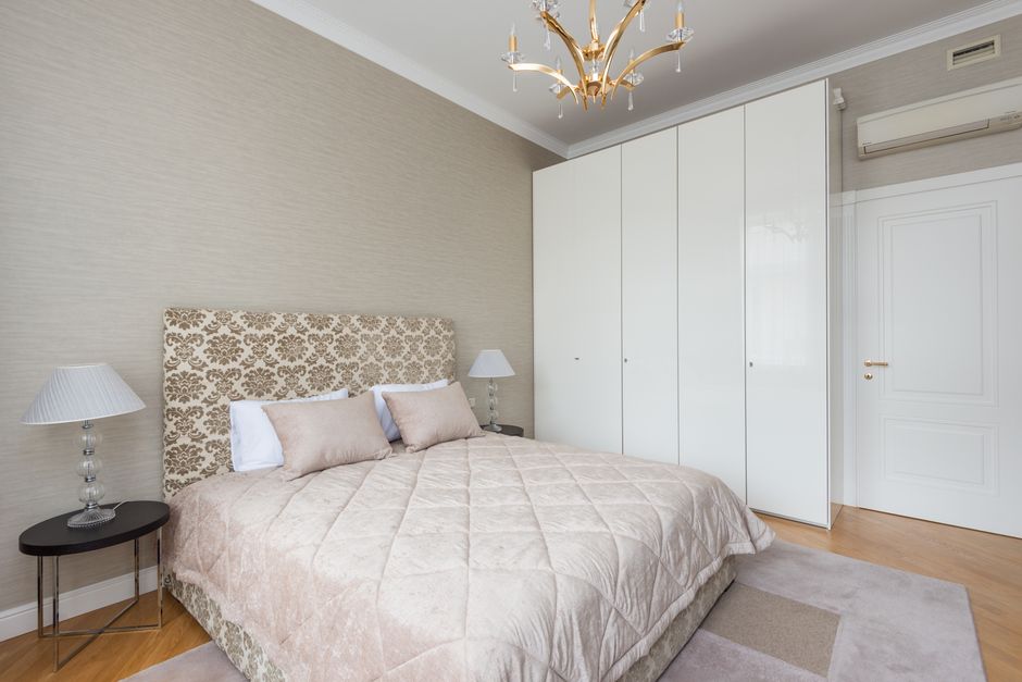 Big bed with ornamental headboard and pillows near lamps on nightstands and glossy closet near door under classic chandelier in bedroom