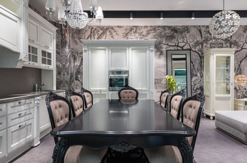 Black table with chairs placed near white cupboards and modern appliances in stylish spacious kitchen with mirror and creative wallpaper